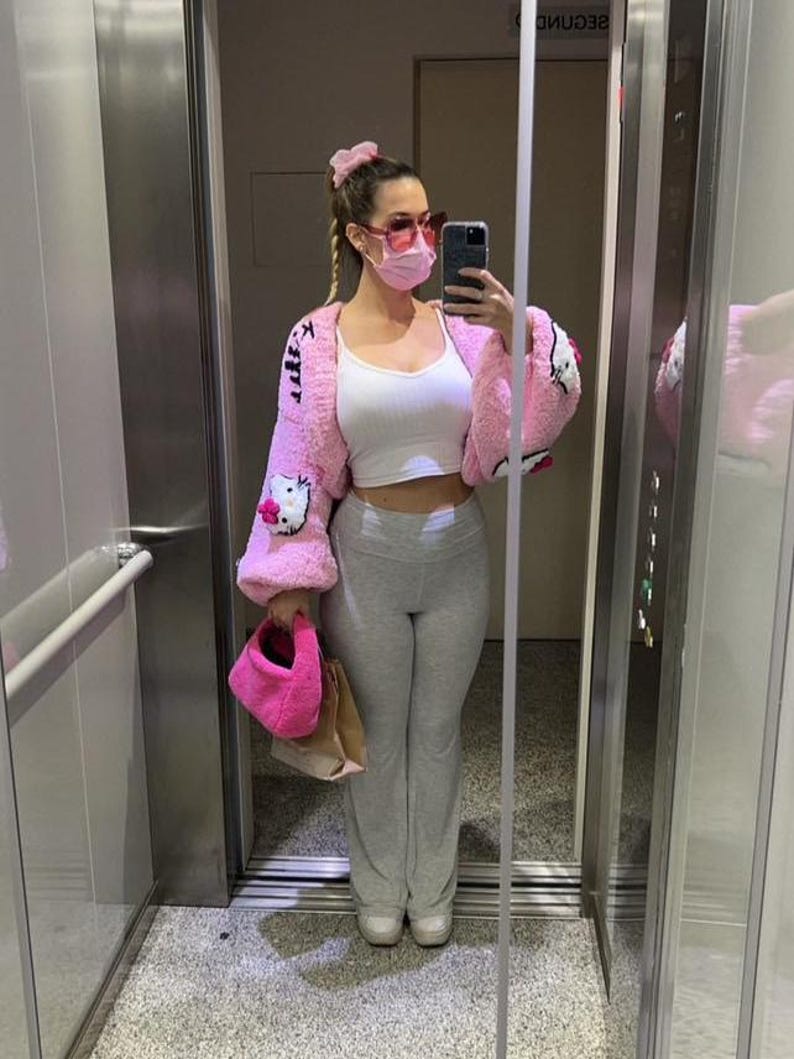 May include: A pink, fuzzy cardigan with Hello Kitty designs, paired with a white top and gray sweatpants. The outfit includes a matching pink purse and scrunchie. The person is wearing pink sunglasses and a face mask.