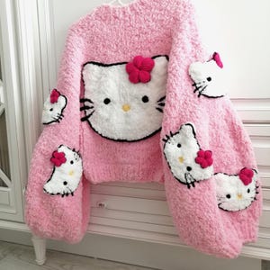 May include: Pink, chunky-knit sweater featuring a large Hello Kitty face on the front and smaller Hello Kitty faces on the sleeves. The sweater has a round neckline and is adorned with red flower accents. The overall design is soft and textured.