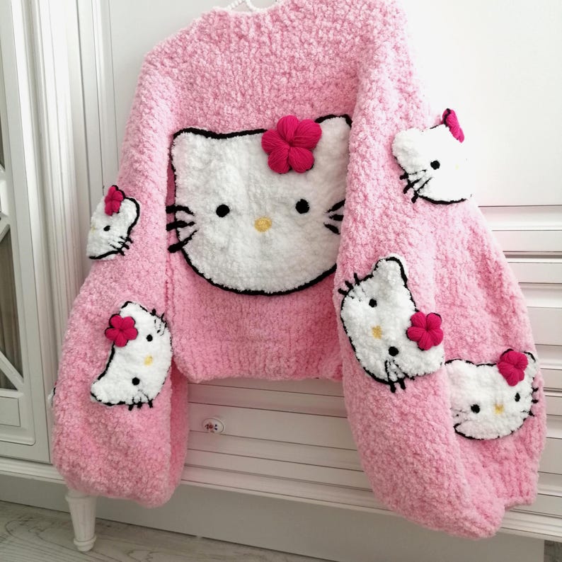 May include: Pink, chunky-knit sweater featuring a large Hello Kitty face on the front and smaller Hello Kitty faces on the sleeves. The sweater has a round neckline and is adorned with red flower accents. The overall design is soft and textured.