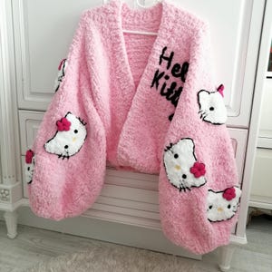 Könnte beinhalten: Ein rosa, flauschiger Cardigan mit langen, voluminösen Ärmeln. Der Cardigan hat mehrere gestickte Hello Kitty-Applikationen mit roten Schleifen. Die Worte "Hello Kitty" sind in Schwarz auf dem oberen Ärmel geschrieben.