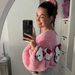 May include: Pink, fluffy, cropped sweater featuring three Hello Kitty face appliqu&eacute;s. The sweater has long sleeves and a round neckline. The Hello Kitty faces are white with black details and a red bow. The sweater is paired with gray leggings.