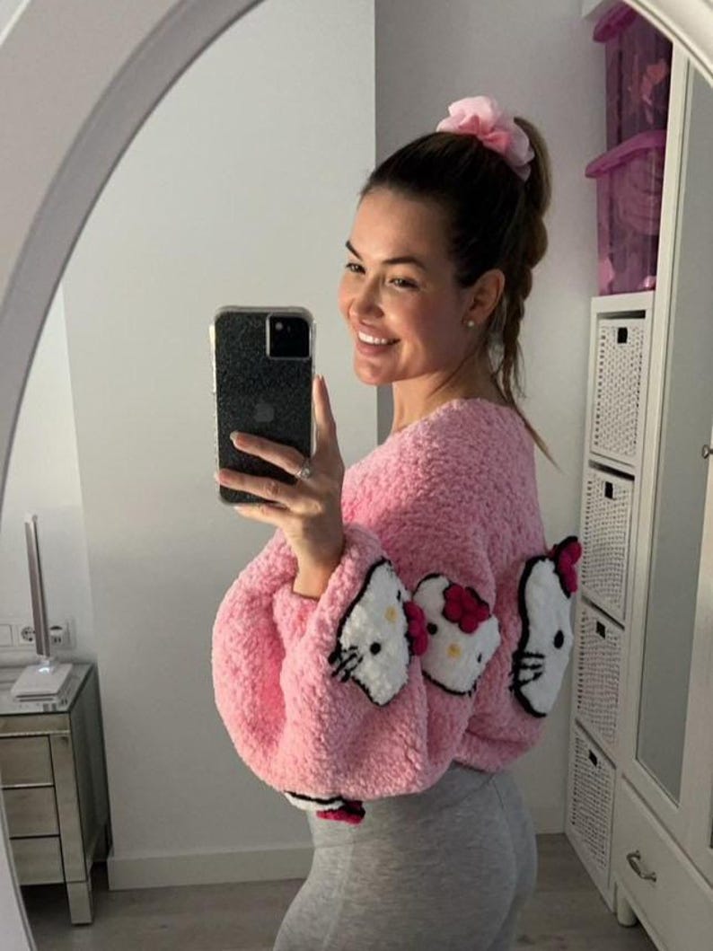 May include: Pink, fluffy, cropped sweater featuring three Hello Kitty face appliqu&eacute;s. The sweater has long sleeves and a round neckline. The Hello Kitty faces are white with black details and a red bow. The sweater is paired with gray leggings.
