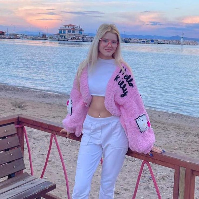 May include: A pink, fluffy cropped jacket with the words "Hello Kitty" in black script on the sleeve. The jacket has a Hello Kitty patch on the front. The person is wearing a white crop top and white pants.