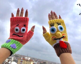 May include: A pair of knitted fingerless gloves, one red and one yellow, each featuring a cartoon character face. The red glove has a green cuff with purple accents, while the yellow glove has a brown cuff and a red tie.