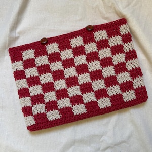 Può includere: Una pochette all'uncinetto rossa e crema con un motivo a scacchiera. La borsa rettangolare ha due bottoni marroni sul bordo superiore. La borsa fatta a mano è su uno sfondo bianco.