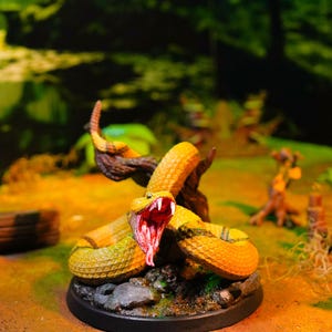 Peut inclure: Miniature détaillée d'un serpent jaune et vert, la gueule ouverte, révélant des crocs et une langue rouges. Le serpent est enroulé autour d'une branche, sur une base noire avec un décor naturel.