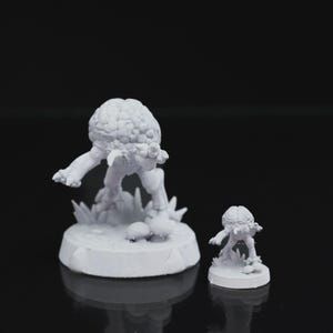 Op de afbeelding: Twee witte miniatuurbeeldjes van een hersenhoofdige creatuur. De grotere figuur staat op een cirkelvormige basis met gras- en paddenstoeldetails. De kleinere figuur weerspiegelt het ontwerp, ook op een cirkelvormige basis. De achtergrond is zwart.