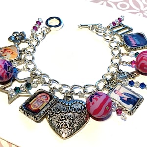 May include: A silver charm bracelet with various charms, including a heart charm with the text "You Rock and Roll", a star charm, a jukebox charm, a flower charm, a charm with the text "Happy Days", a charm with a picture of a person, and several pink and purple beads.