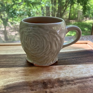 May include: A ceramic mug with a light gray glaze and a swirling, textured design. The mug has a rounded body and a handle.