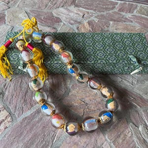 May include: A beaded necklace featuring clear, oval-shaped beads with miniature painted faces and gold accents. The necklace has yellow tassels and is displayed on a patterned green fabric.