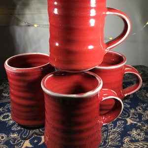 May include: Four red ceramic mugs with white rims. The mugs have a textured, slightly rippled surface. They are stacked on top of each other, with the top mug slightly tilted.