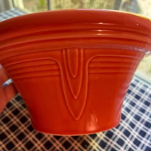 May include: A vibrant orange ceramic bowl with a distinctive Art Deco design. The bowl features horizontal ridges around the rim and a central, stylized vertical design. The bowl is set against a checkered blue and white tablecloth.