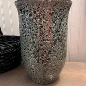 May include: A tall, speckled vase with a flared top. The vase has a dark gray base color with black and white speckles. The vase is made of ceramic and has a smooth, glossy finish. The vase is a decorative item.