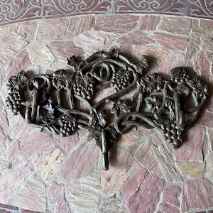 May include: Ornate bronze-colored wall hook featuring a grape vine design. The hook is detailed with clusters of grapes and leaves, adding a touch of elegance. The decorative piece is mounted on a textured, multi-colored stone surface.
