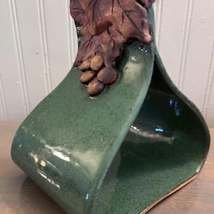 May include: A handcrafted, green ceramic basket with a leaf and grape cluster design. The basket has a curved, open shape with a gold rim. The leaf and grape cluster are brown and add a naturalistic touch to the piece.