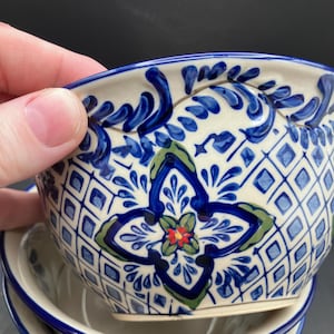 May include: A stack of ceramic bowls with a white base and intricate blue floral patterns. The bowls feature a diamond grid design and a central floral motif with green and red accents. The rim is adorned with a blue wave pattern.