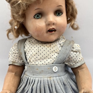 May include: Vintage doll with blonde curly hair and green eyes. The doll wears a light blue dress with a white polka dot top. The doll's face shows signs of age, with visible cracks.