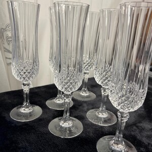 May include: A set of five clear glass champagne flutes featuring a tall, slender bowl with vertical fluting and a textured diamond-cut pattern at the base. Each flute stands on a clear glass stem and a circular base, showcasing a reflective finish.