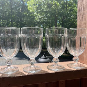 May include: Four clear, stemmed water goblets with vertical ribbed detailing. The glasses are displayed on a wooden surface with a blurred green background of trees and sunlight.