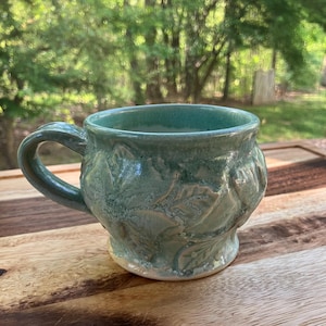 May include: A green ceramic mug with a textured leaf design. The mug has a rounded body and a handle.