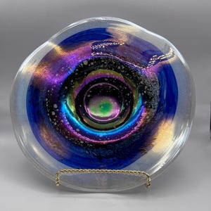 May include: A clear glass bowl with a swirling, iridescent design. The center features a vibrant mix of colors, including green, purple, and blue, surrounded by concentric rings of color. The bowl is supported by a gold-colored metal stand.
