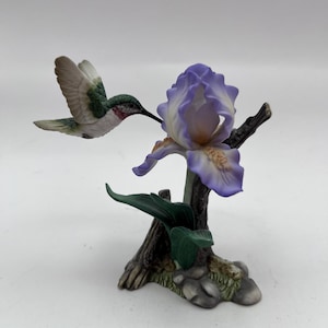 May include: A ceramic figurine of a hummingbird hovering near a purple and white iris flower. The hummingbird has green, white, and red details. The flower and bird are perched on a brown branch with green leaves and a rocky base.
