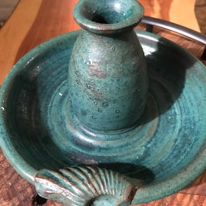 May include: A teal blue ceramic incense burner with a seashell handle. The burner has a small, round opening at the top and a larger, round base.
