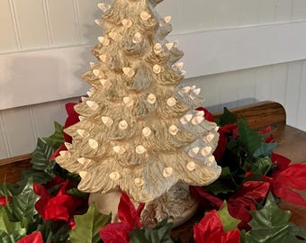 May include: A ceramic Christmas tree with built-in lights, painted in a beige color. The tree is surrounded by a garland of red poinsettias and green leaves, creating a festive holiday display.