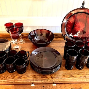 May include: A collection of ruby red glassware and dishware, including wine glasses, tumblers, mugs, plates, a bowl, and a serving tray. The items are arranged on a wooden surface, showcasing a vintage aesthetic. The glassware features intricate patterns.