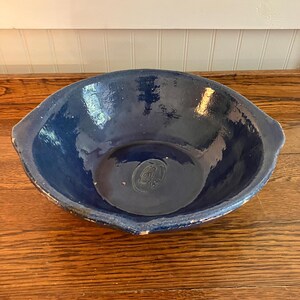 May include: A large, deep blue ceramic bowl with a glossy finish. The bowl has a wide, oval shape with slightly raised edges and small handles. The interior features a subtle embossed design. The bowl is set on a wooden surface.
