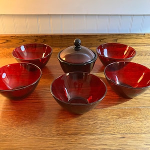 May include: A set of six red-toned wooden bowls and a lidded serving dish. The bowls have a glossy finish and a dark rim. The serving dish has a matching dark lid with a small, round handle. The items are arranged on a wooden surface.