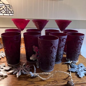 May include: A collection of ruby-colored glassware, including martini glasses and tall tumblers. The tumblers feature an etched design. Decorative snowflake ornaments and a bell are also present, suggesting a holiday theme.