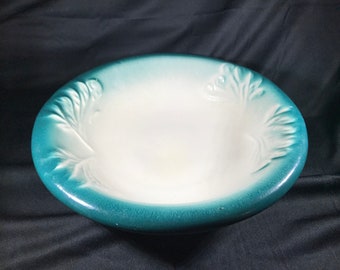 May include: A white ceramic bowl with a teal green rim and embossed floral designs. The bowl is slightly oval shaped.
