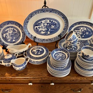 May include: A collection of blue and white ceramic tableware, including plates, bowls, cups, saucers, and serving dishes. The pieces feature a classic Willow pattern design. The set is arranged on a wooden surface.