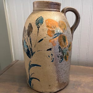 May include: A tan stoneware jug with a handle and a wide mouth. The jug is decorated with hand-painted flowers in orange, blue, and brown. The jug has a rustic, vintage appearance.