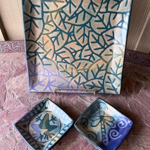 May include: A square ceramic serving platter and two small square dishes. The platter has a teal leaf pattern on a beige background. The small dishes feature a frog and a dragonfly design, respectively. The color palette includes teal, blue, and purple.