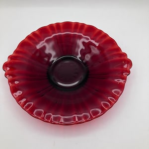 May include: A ruby red glass bowl with a ruffled edge and a circular center. The bowl has a deep, rich color and a glossy finish. The ruffled edge adds a decorative touch to the bowl.