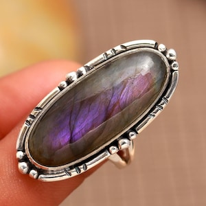 Puede incluir: Un anillo de plata con una gran piedra ovalada de labradorita. La piedra tiene una base gris oscura con destellos iridiscentes púrpuras y azules.