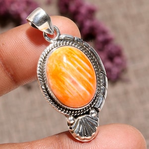 May include: Oval pendant featuring an orange and white stone set in a silver-coloured metal frame. The pendant has a bail for a chain and a shell-like design at the bottom. The stone has a striped pattern.