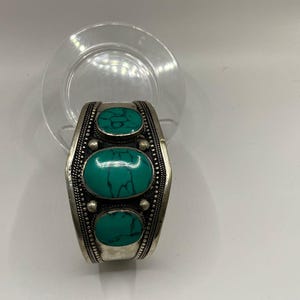 May include: Silver cuff bracelet with three oval turquoise stones set in the center. The bracelet has a beaded design around the stones and along the edges.