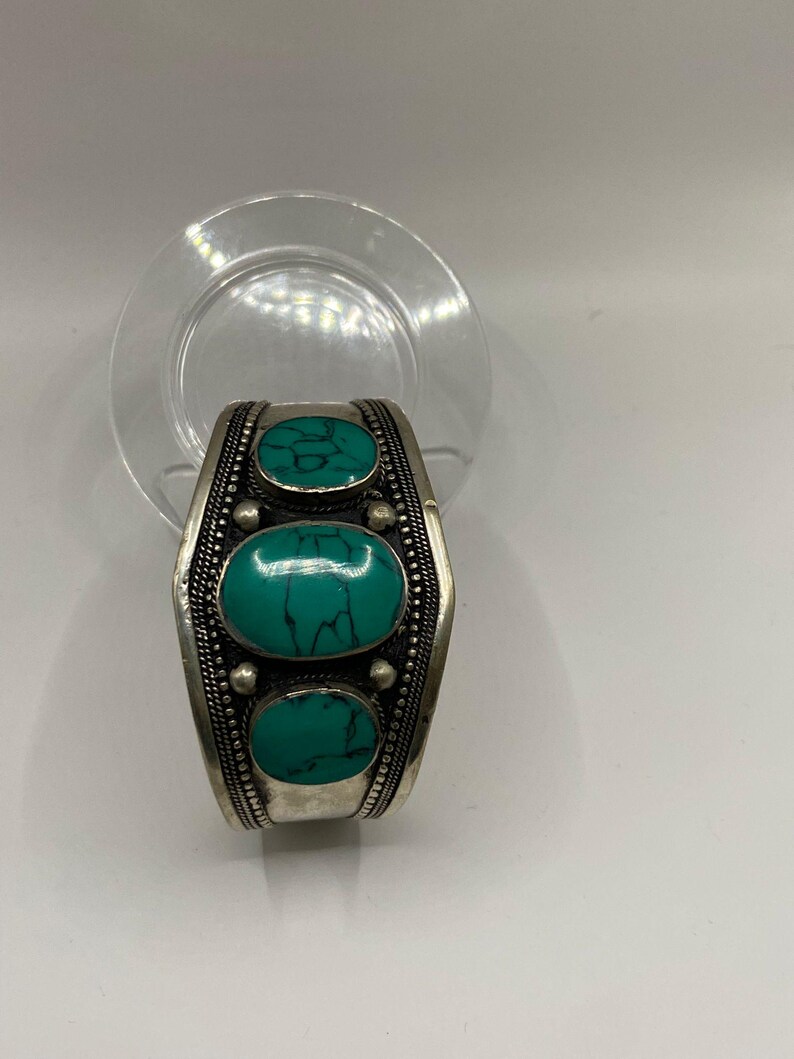 May include: Silver cuff bracelet with three oval turquoise stones set in the center. The bracelet has a beaded design around the stones and along the edges.