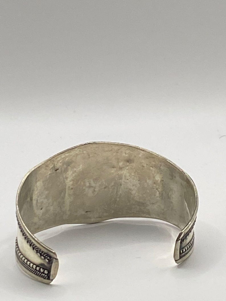 May include: Silver cuff bracelet with a textured design and a decorative border.