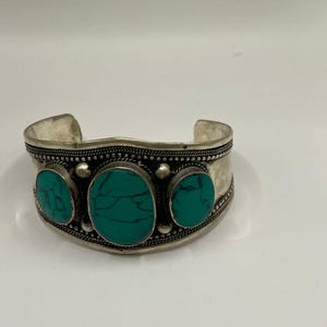 May include: A silver cuff bracelet with three turquoise stones set in the center. The bracelet has a decorative design around the edge.