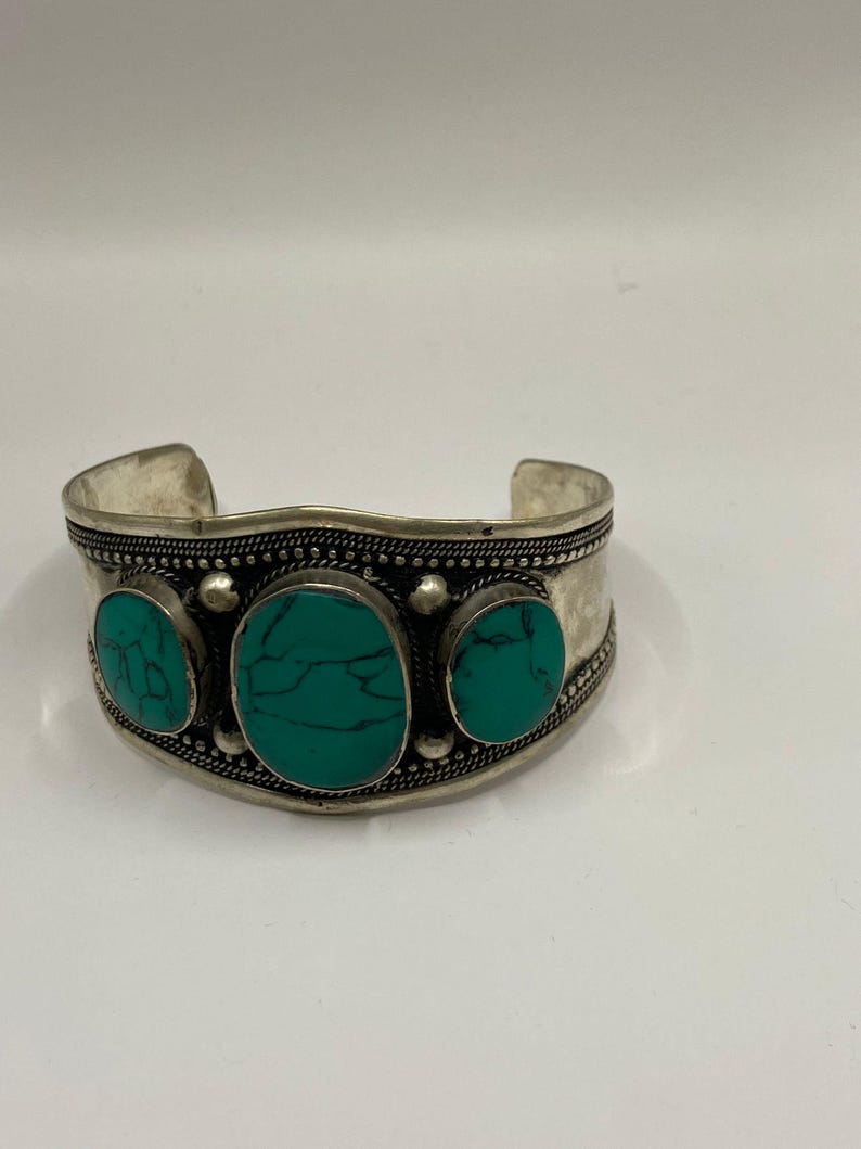 May include: A silver cuff bracelet with three turquoise stones set in the center. The bracelet has a decorative design around the edge.