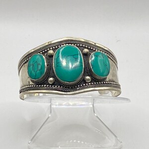 May include: Silver cuff bracelet with three turquoise stones set in the center. The bracelet has a beaded design around the edge.