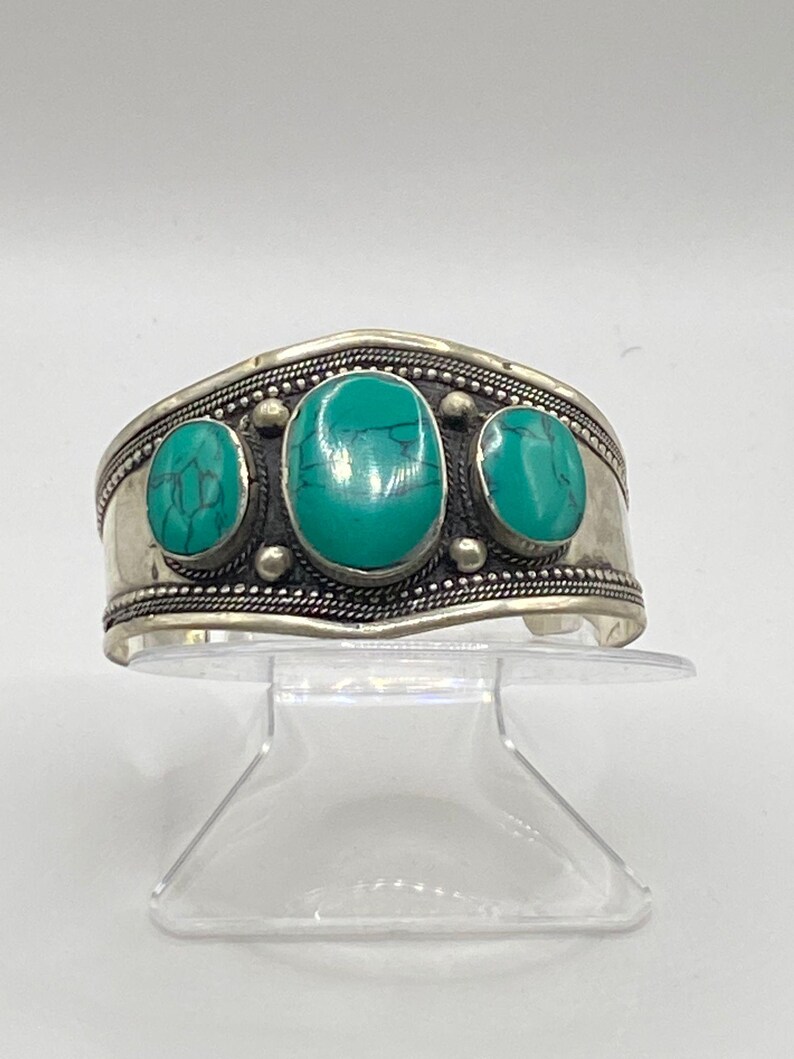 May include: Silver cuff bracelet with three turquoise stones set in the center. The bracelet has a beaded design around the edge.