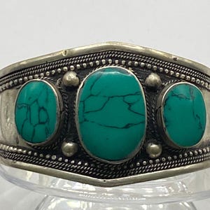 May include: A silver bracelet with three turquoise stones set in the center. The bracelet has a decorative border around the edge.