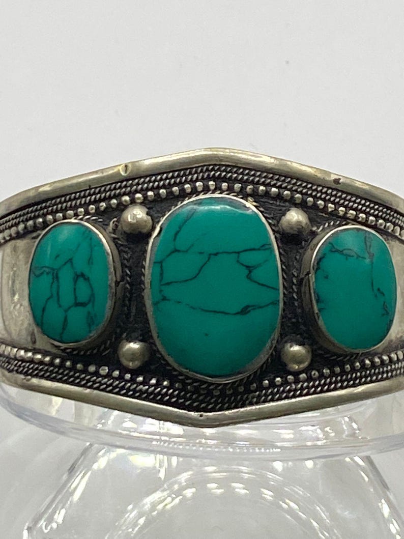 May include: A silver bracelet with three turquoise stones set in the center. The bracelet has a decorative border around the edge.