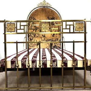 Può includere: Un letto antico in ottone con testiera e pediera decorative. La struttura del letto presenta dettagli ornati e un materasso a righe con un motivo floreale. La testiera ha un pannello decorativo con un'immagine vintage.