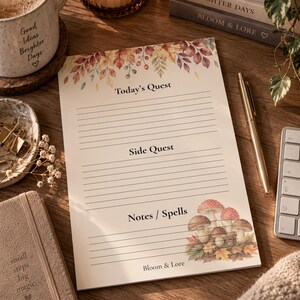 Peut inclure: Un bloc-notes rectangulaire en papier orné d'une bordure de feuilles d'automne en haut et d'une illustration de champignons en bas à droite. La page comporte les titres Today's Quest, Side Quest et Notes / Spells, avec le texte Bloom & Lore.
